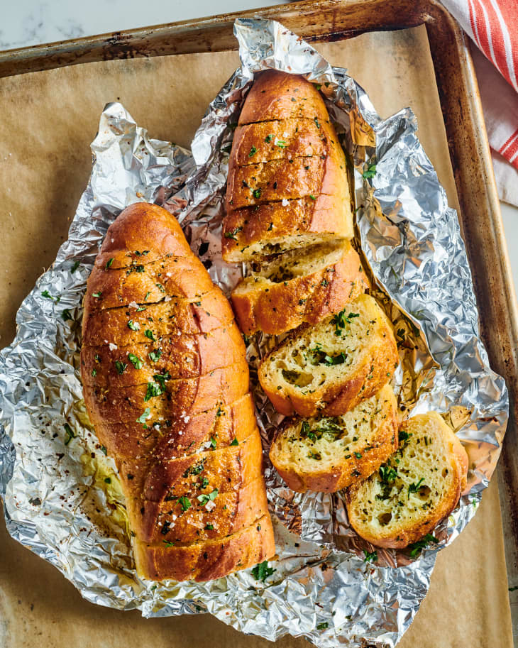 slices of the garlic bread sit next to a garnished, scored loaf in foil