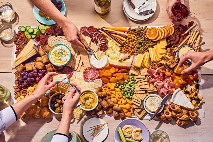grazing table or board on a light wood table. There is meat, cheese, crackers, fruit, vegetables, dips, olives, and more. Plates and glasses, toothpicks on the table by the food. Hands are reaching in to get snacks.