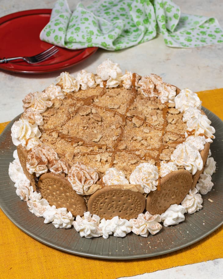 Angled-down photo of a coffee icebox cake decorate with whipped cream and cookies on platter. Gold runner underneath. Red plate with fork and green and white napkin in background