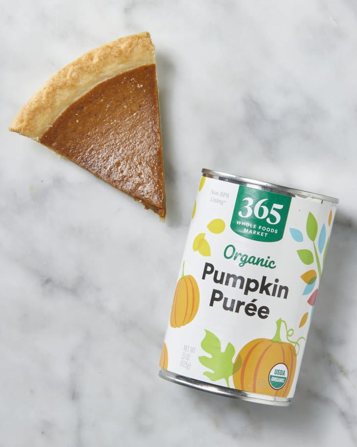 Can of 365 Organic Pumpkin Puree with a slice of pumpkin pie next to it on a marble surface.