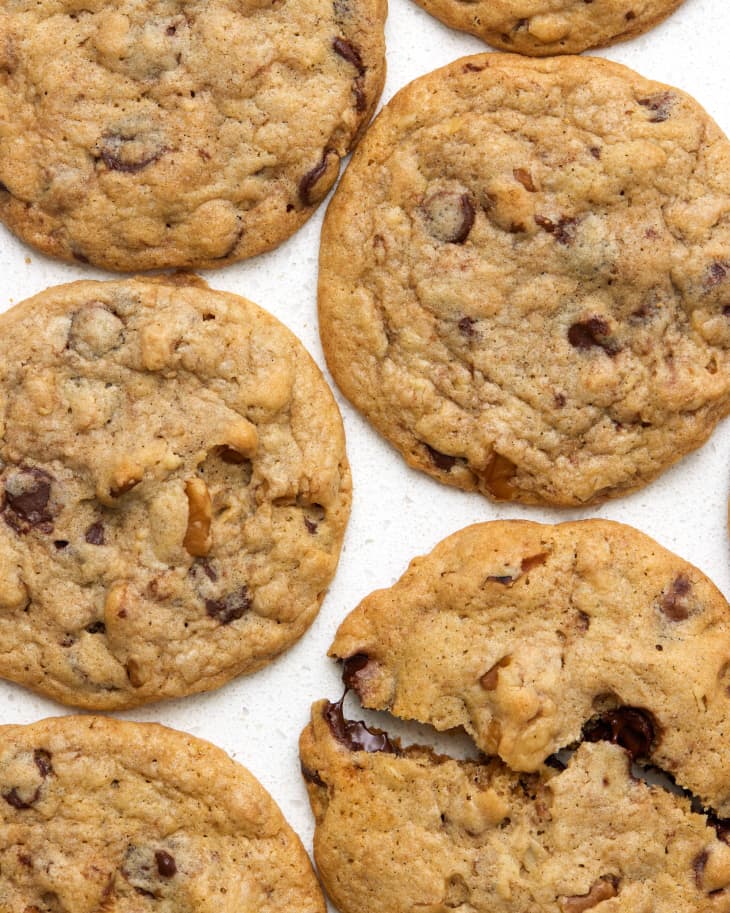 Overhead view of chocolate chip cookies on a white stone surface and the cookie in the bottom right corner is cut in half.