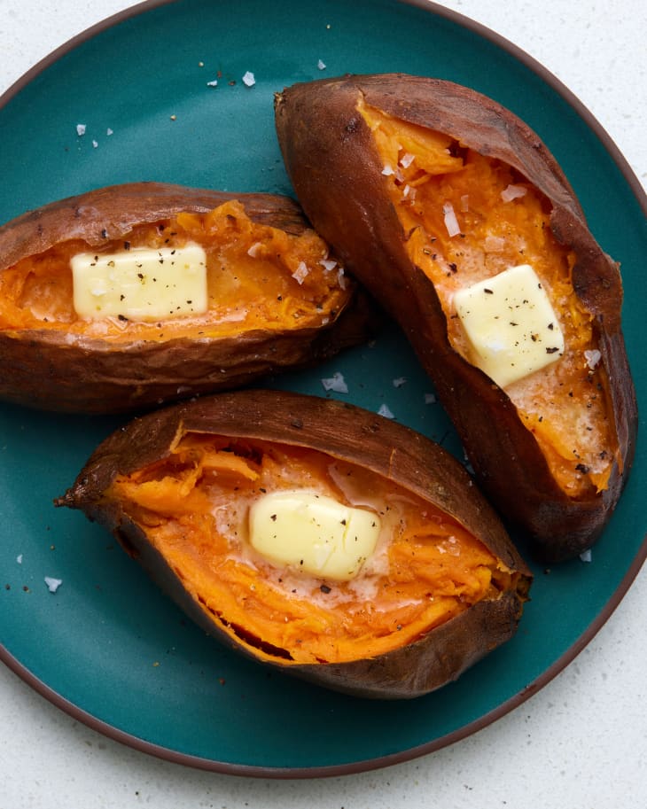 Overhead view of 3 baked sweet potatoes cut open on a dark blue plate, topped with butter and black pepper.
