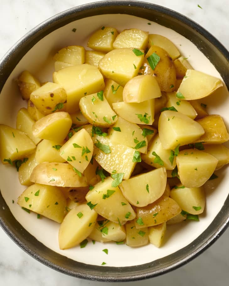 Overhead view of boiled potatoes in a white bowl with a brown rim, topped with herbs.