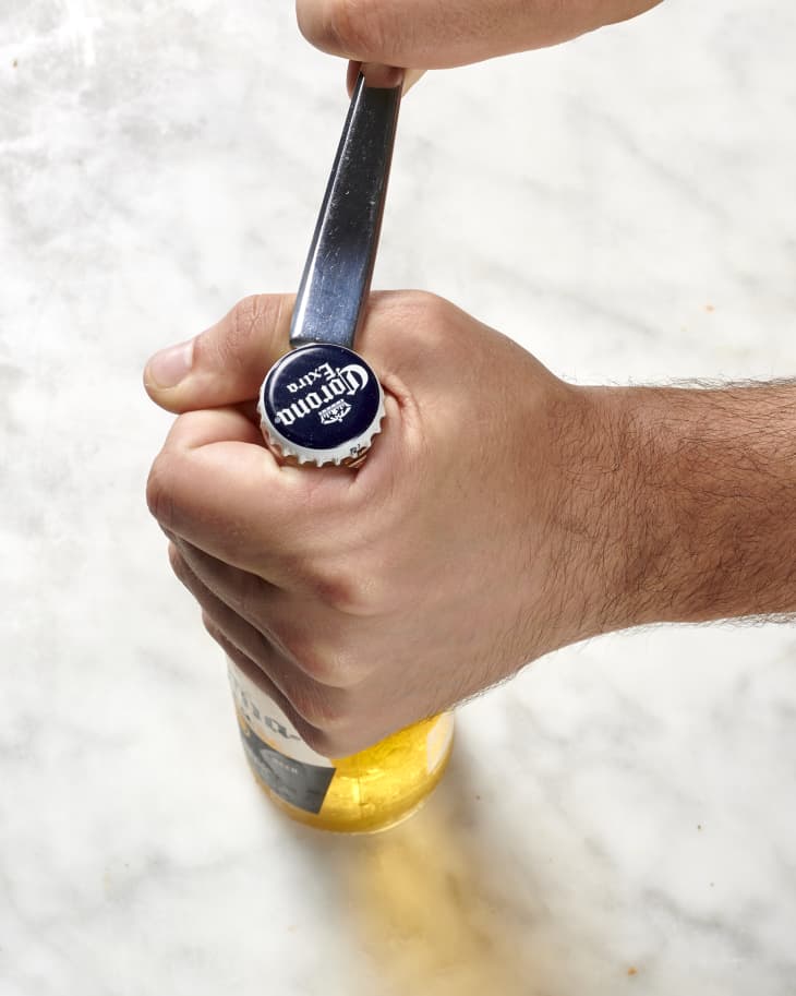 Overhead view of one hand holding the neck of a beer bottle while the other hand pries open the lid with the back end of a spoon.