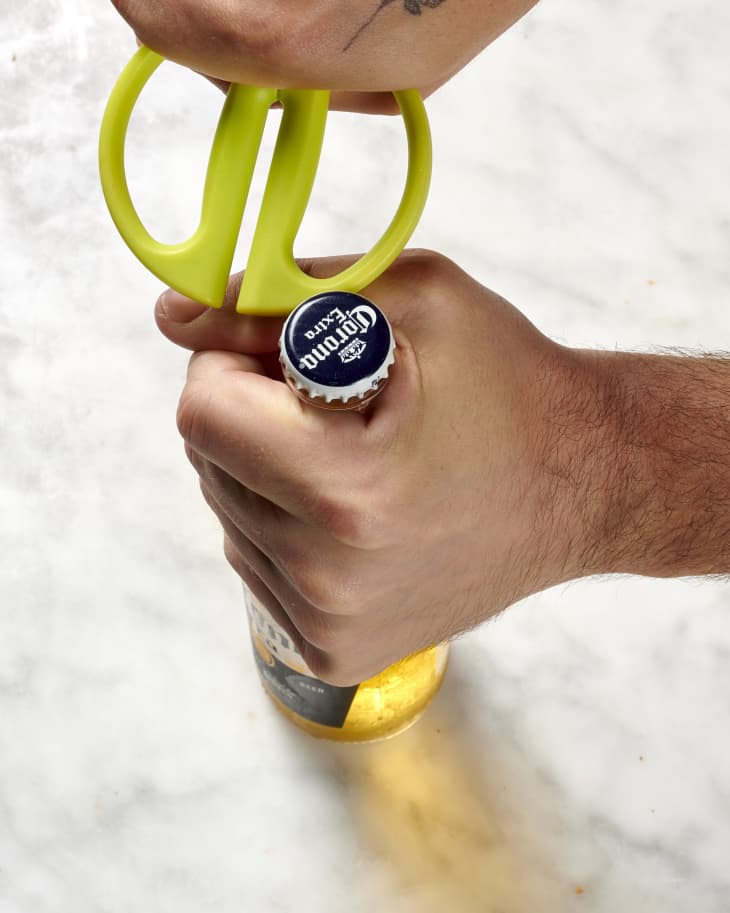Overhead view of one hand holding the neck of a beer bottle, while the other hand opens the lid with the handle of green scissors.