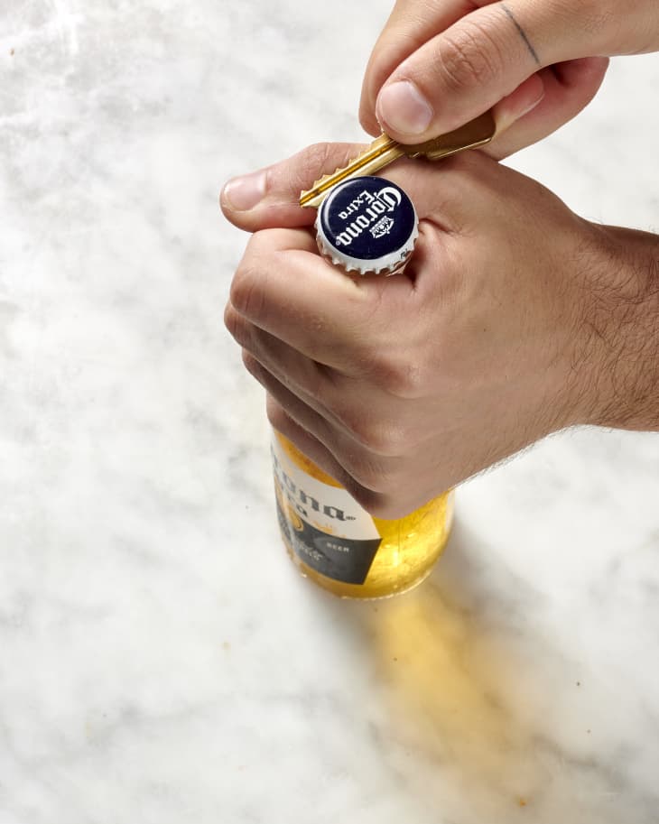 Overhead view of one hand holding the neck of a beer bottle while the other hand opens the lid with the flat edge of a key.