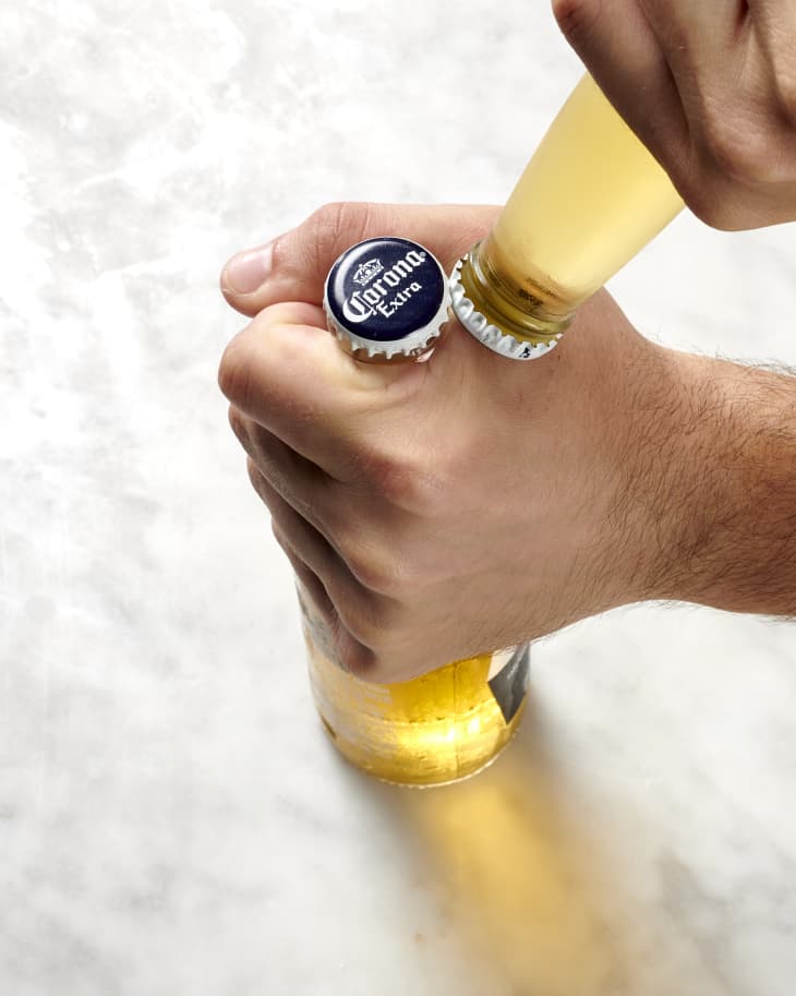 Overhead view of on hand holding the neck of a beer bottle, while the other hand holds another beer bottle upside down, with both bottle lids touching on the side.