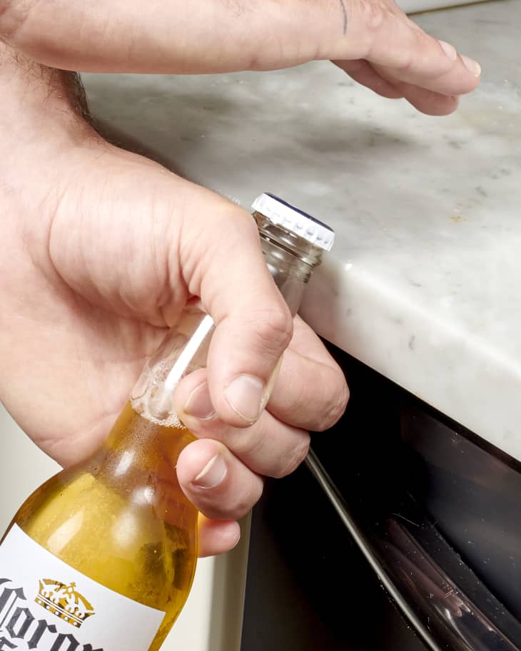 Side view of a hand holding the neck of a beer bottle and holding the lid against the edge of a marble countertop.