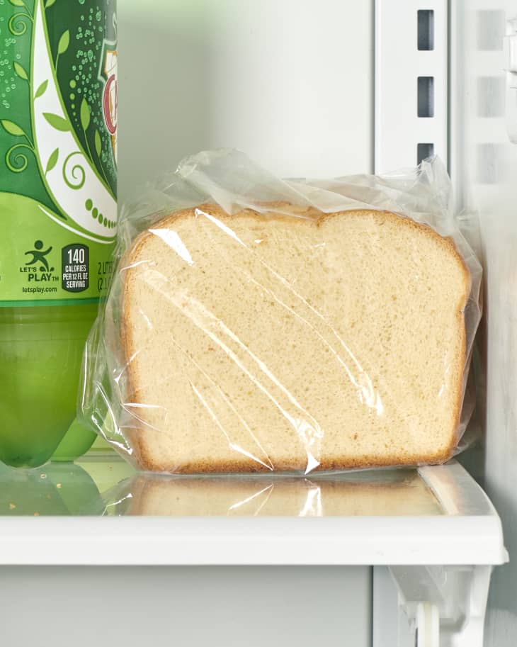 Front view of white bread wrapped in plastic on a shelf in the refrigerator, next to a green bottle of ginger ale.