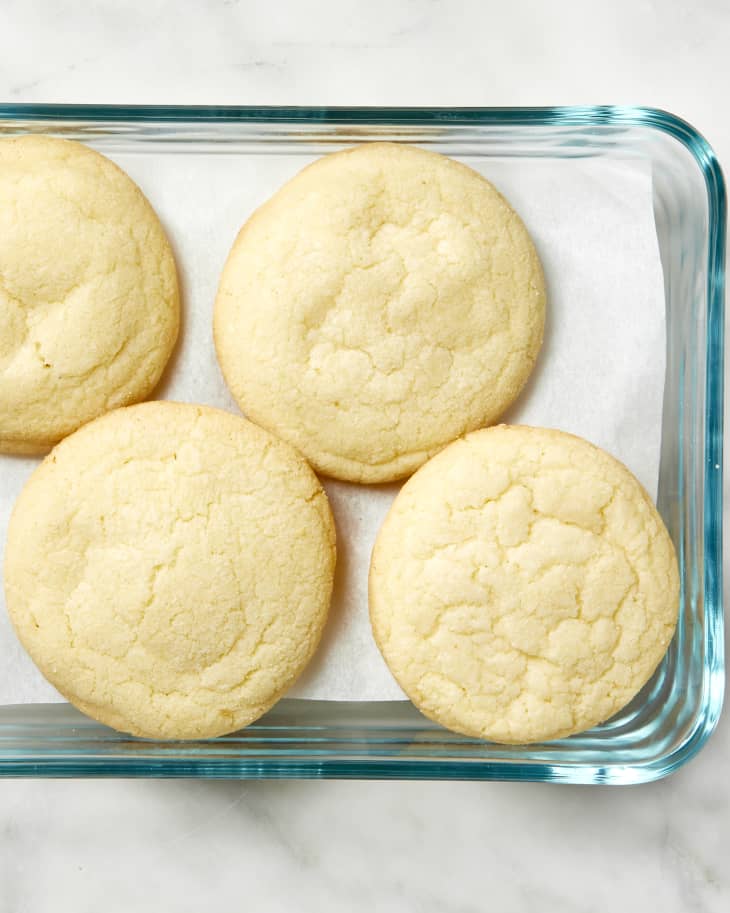 Close up overhead shot of sugar cookies being stored in a glass container, with parchment paper at the bottom.