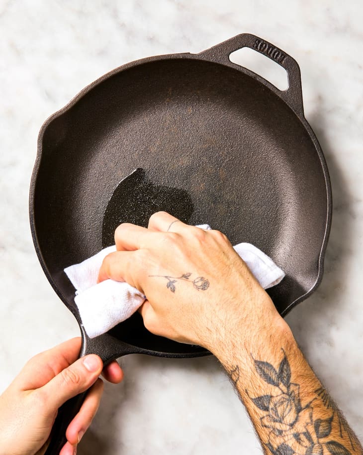 A hand wiping a cast iron skillet with neutral oil with a dish towel.