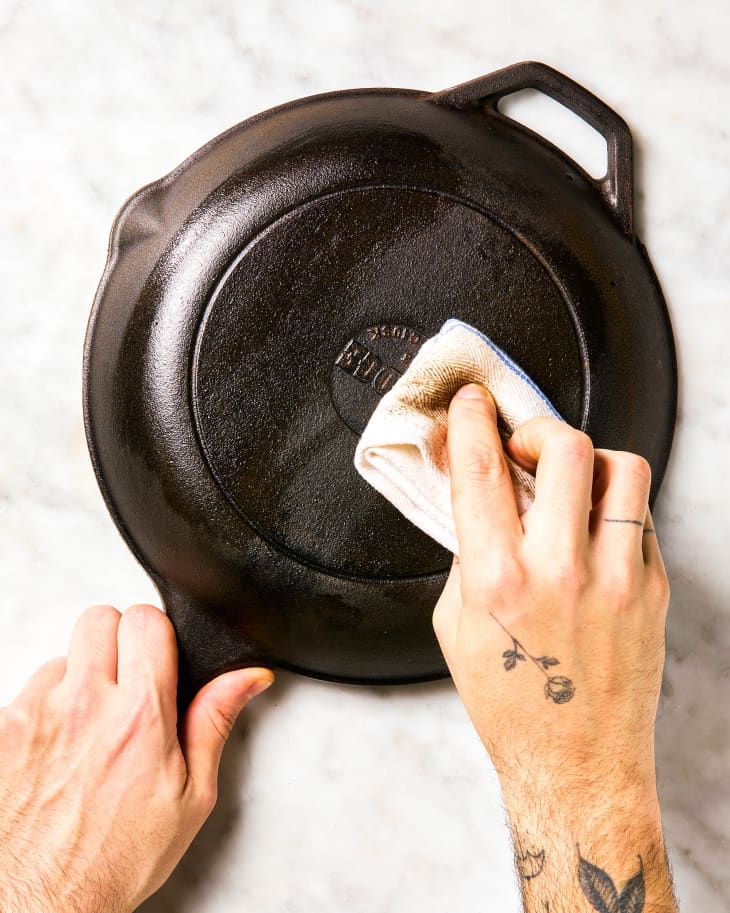 A hand wiping a cast iron skillet with neutral oil with a dish towel.