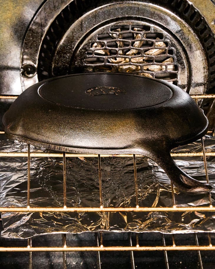 A cast iron skillet in an oven, upside down on a rack with aluminum foil underneath to catch dripping oil.