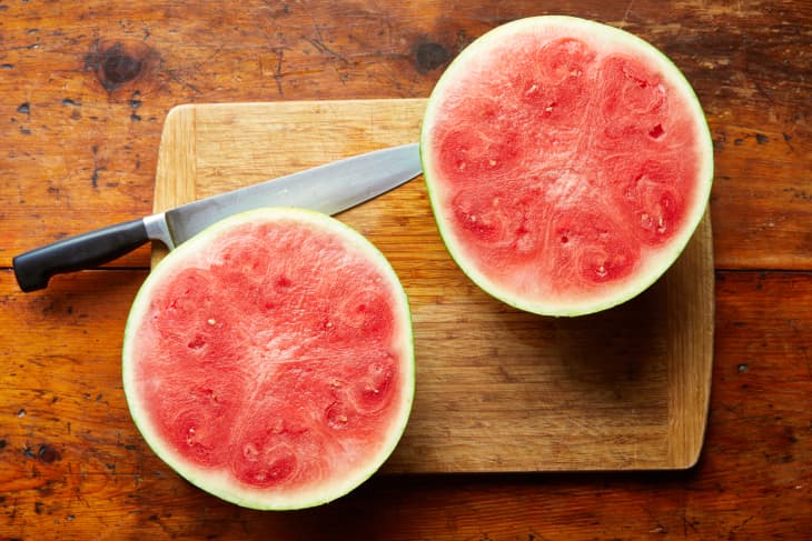 A halved watermelon sits on a cutting board with a chefs knife.