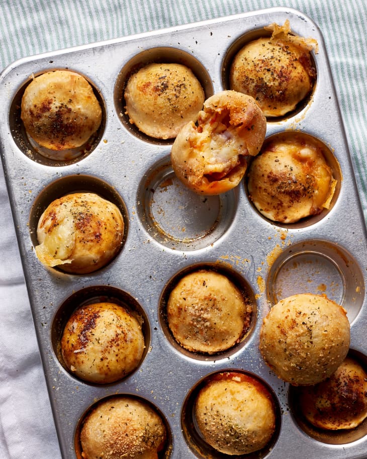 Pizza bombs baked in crust to a rich golden-brown hue; a bread lays on top of a muffin tin to show its melted cheese filling