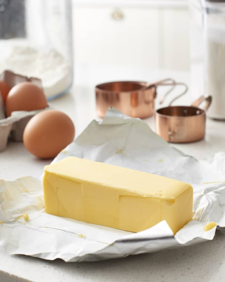 A stick of butter on its open wrapping with eggs and measuring cups in the background