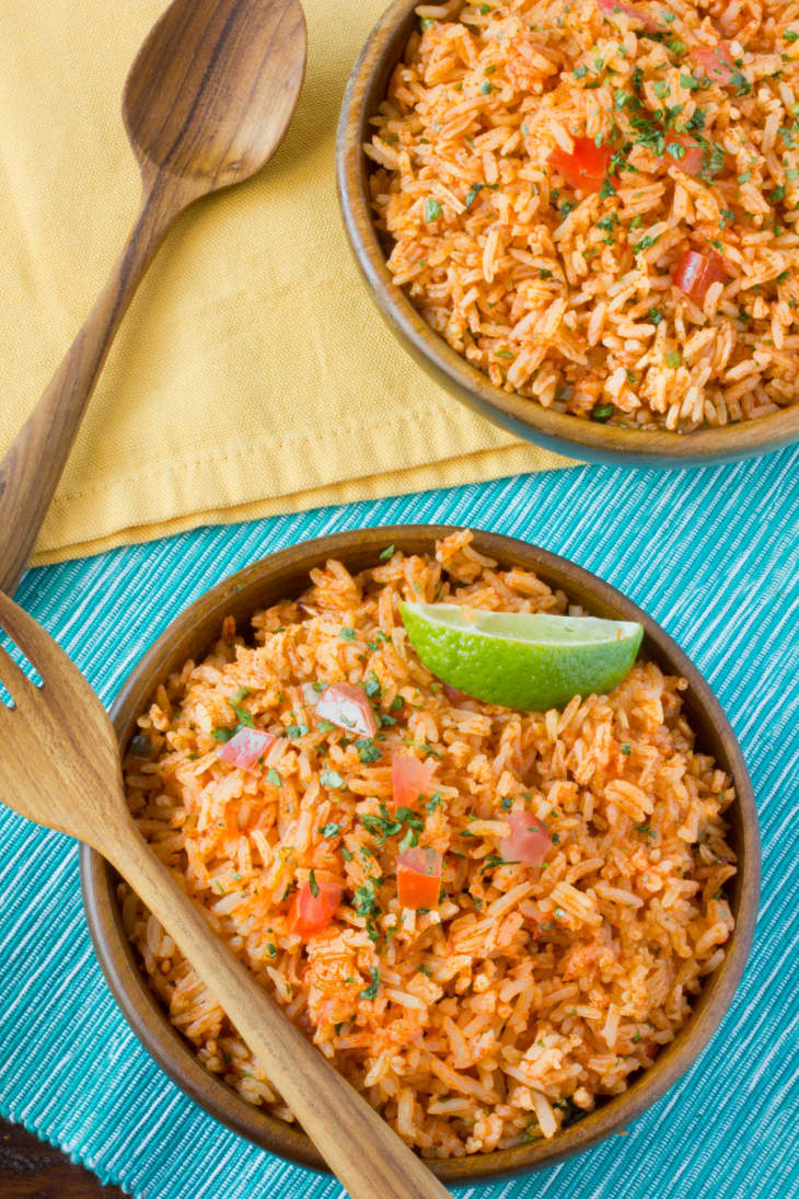 Fluffy Mexican rice, garnished with chopped tomatoes, cilantro, and a wedge of lime, in two separate wooden bowls. A bowl comes with a wooden spoon on the side, while the other with a wooden fork on top
