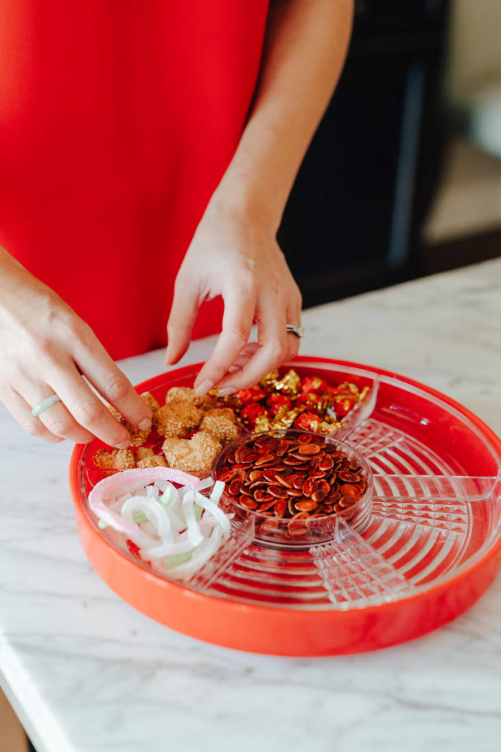 Person arranging snacks in a red tray with seeds, candies, and sliced onions on a marble countertop.