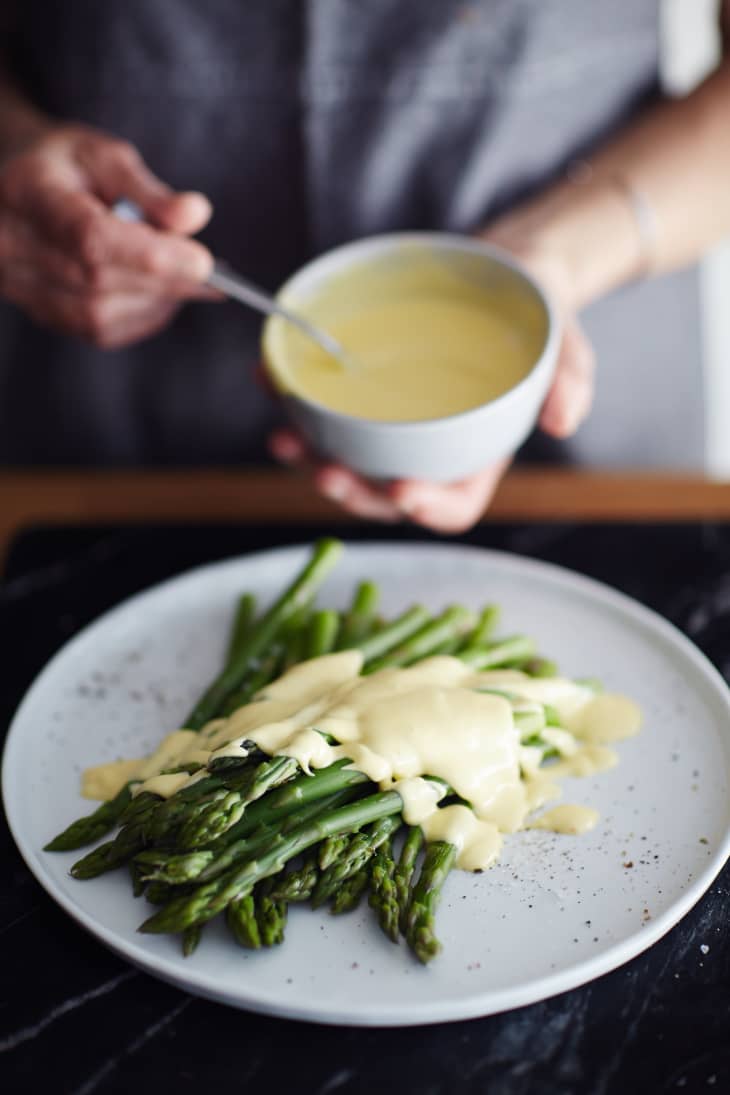A cook holds a small white bowl filled with Hollandaise sauce and a plate of green asparagus topped with Hollandaise sauce