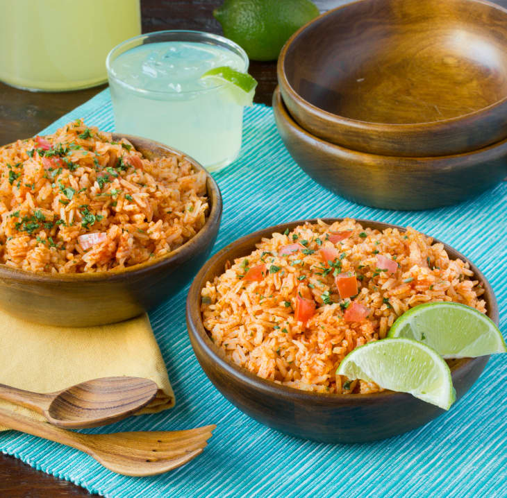 Two bowls of Mexican rice, topped with chopped tomatoes and garnished with cilantro and lime wedges, are placed on an aqua blue table mat