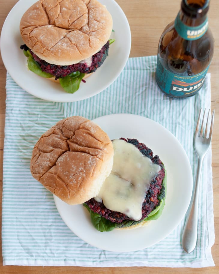 A top shot of two veggie burgers, with melted cheese and lettuce between burger buns, on a plate with a fork on the side, and a bottle of beer