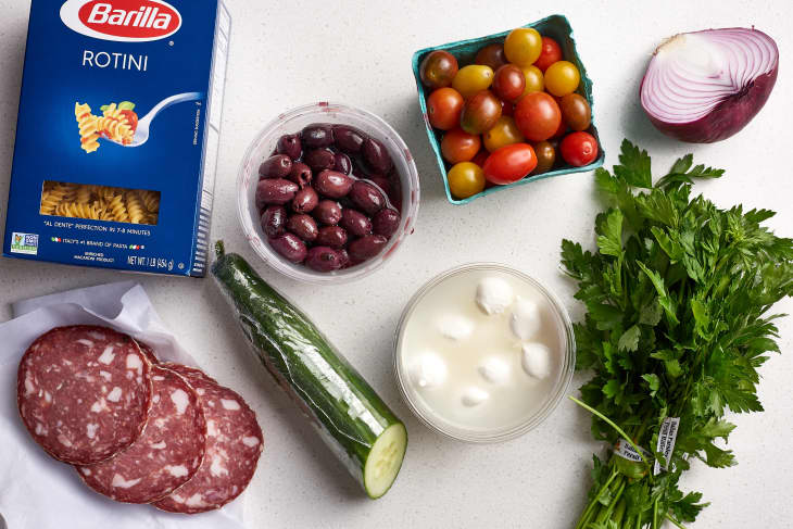 Ingredients for pasta salad, including a box of rotini, English cucumber, red onion, cherry tomatoes, kalamata olives, parsley, salami slices, and mini mozzarella balls