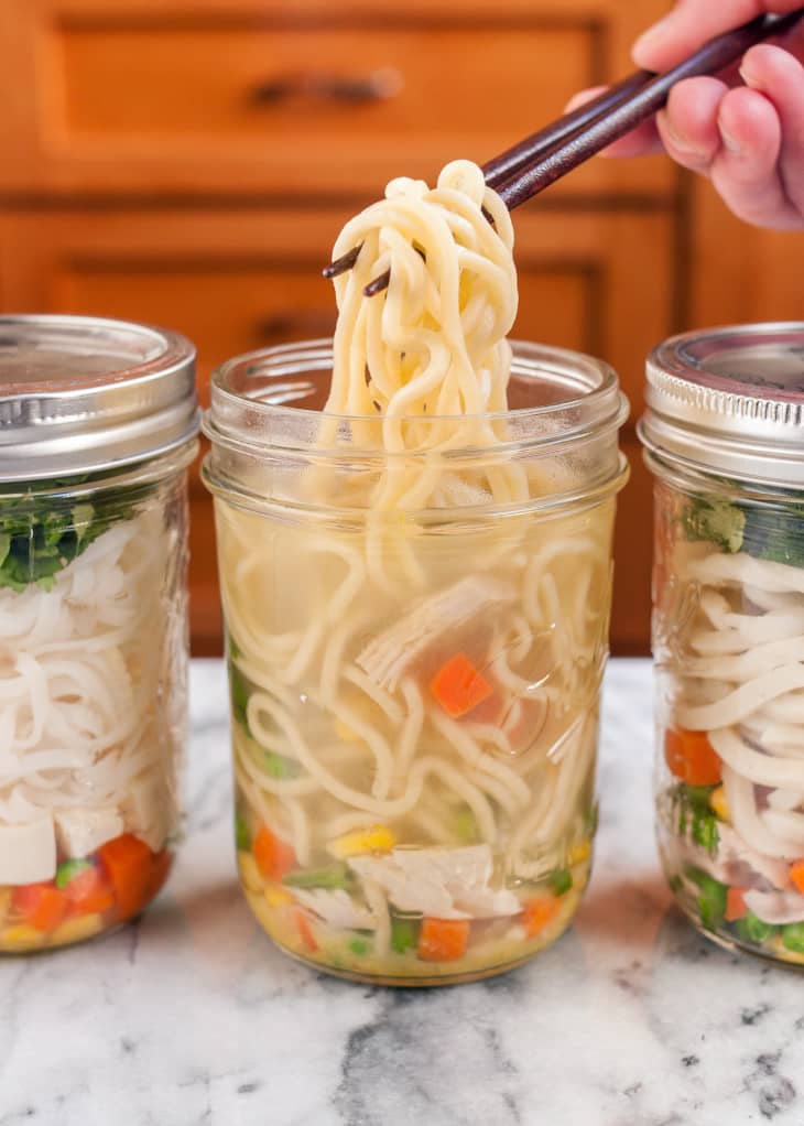 Someone uses chopsticks to get noodles from a mason jar filled with soup stock, carrots, tofu, and peas