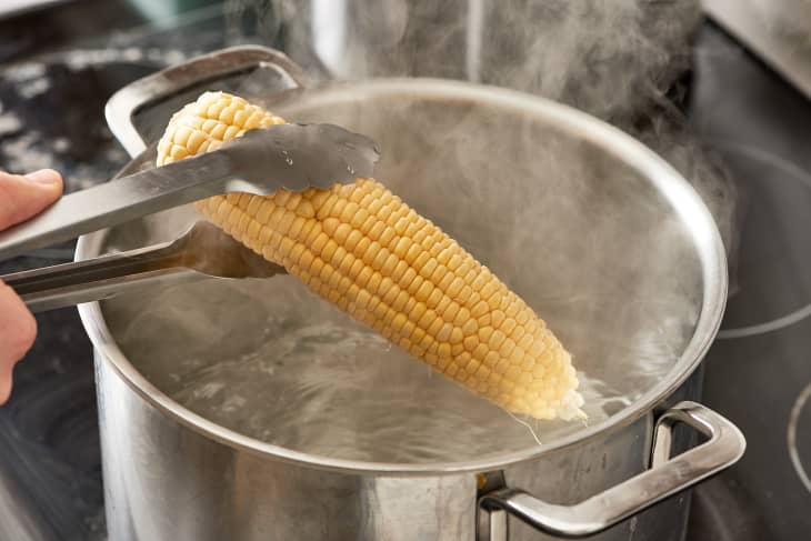 Someone picks a corn using a tong and is about to drop into a pot of boiling water