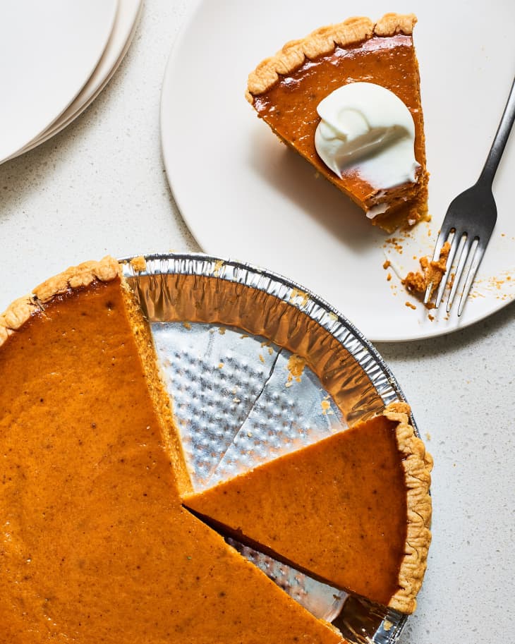 A pumpkin pie in the pan with a slice cut out and served on a plate with whipped cream