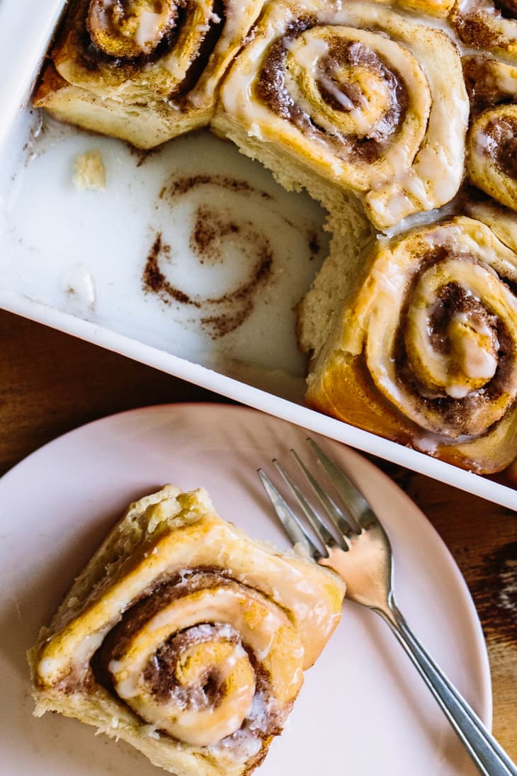 A cinnamon roll on a small plate and beside it is a baking dish filled with freshly baked cinnamon rolls