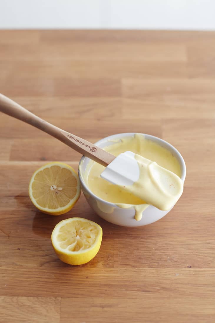 A wooden spatula is placed on top of a white small bowl filled with Hollandaise sauce, and a lemon cut into half is placed beside the bowl