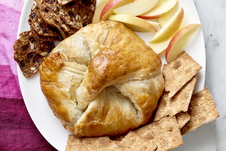 Baked brie in puff pastry surrounded by thinly sliced crisp breads, apple slices, and sesame crackers