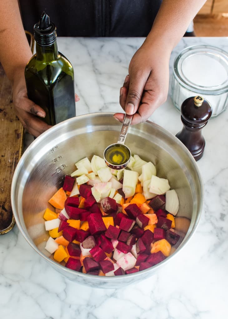 Large metal mixing bowl filled with chopped vegetables including sweet potatoes, beets, and potatoes; a hand holds a tablespoon of olive oil over the bowl, ready to pour in the oil
