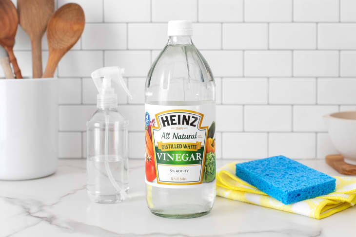 A bottle of Heinz distilled white vinegar on a kitchen counter next to sponges, a cleaning cloth, and a spray bottle