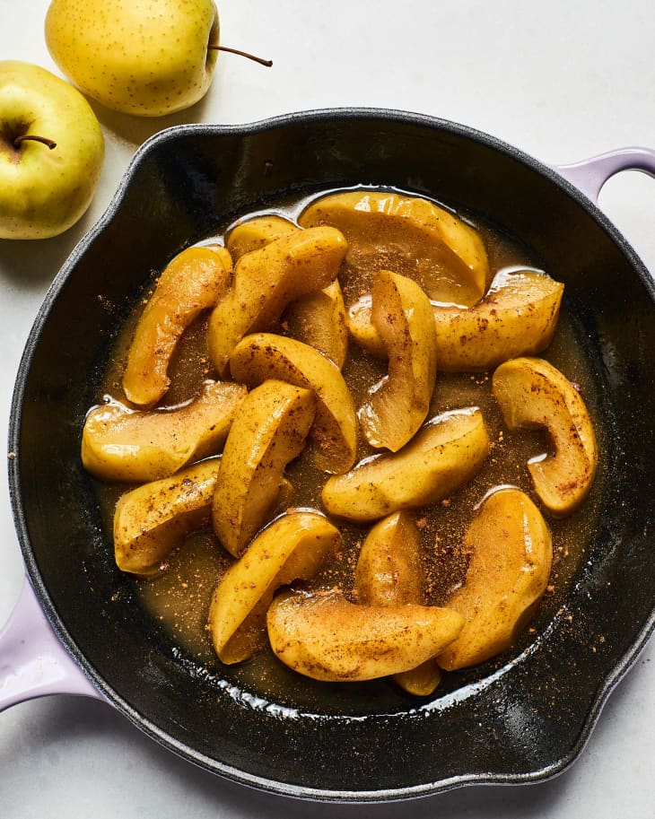 Sliced cooked apples in cast iron skillet in a glaze with ground spices and whole apples in background.
