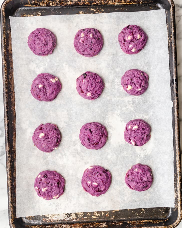 Overhead photo of purple-hued blueberry cookies on baking sheet and white parchment