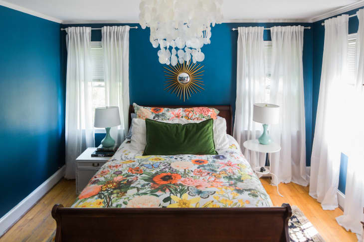 A bedroom with a white chandelier, royal blue walls, a midcentury modern mirror and a floral bedspread.