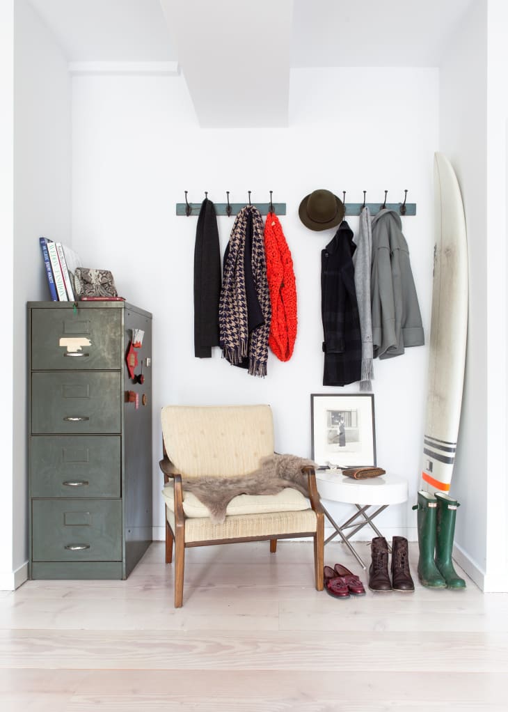 A simple entryway with a metal filing cabinet, coat hooks on the wall, a vintage chair, and a surfboard against the wall