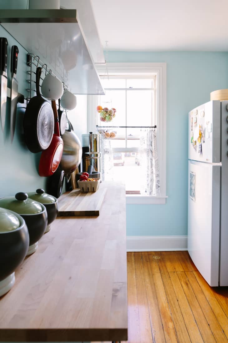 A narrow kitchen with pans on the wall and a window in the background