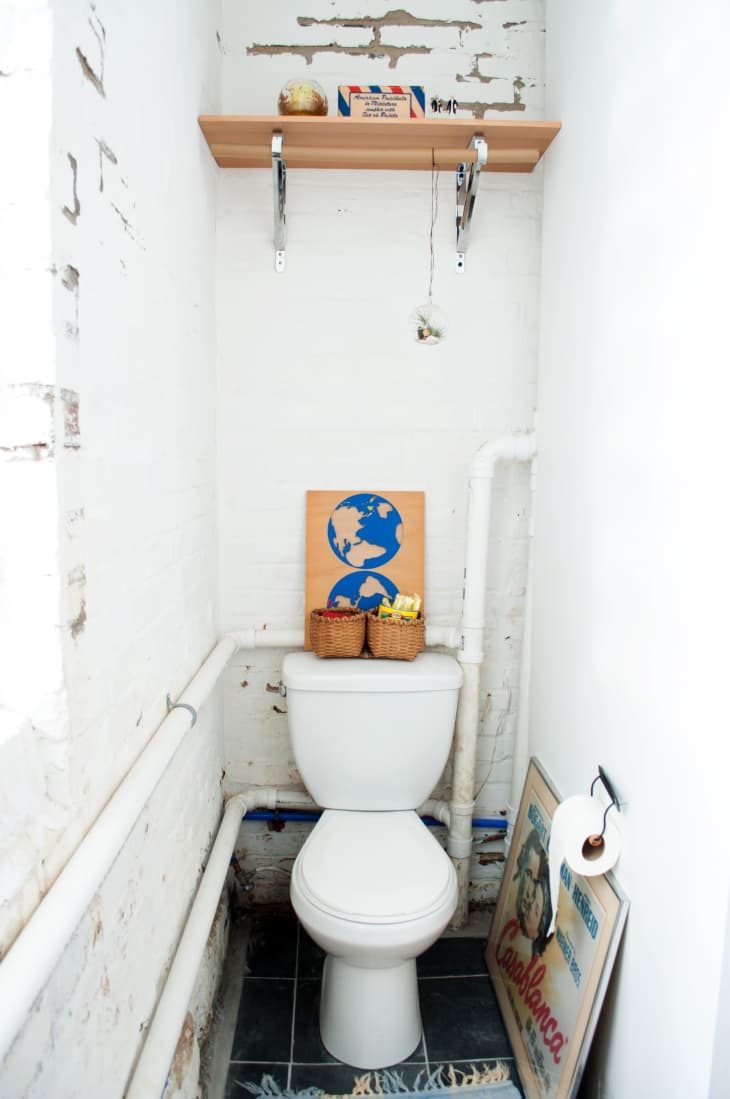 A skinny bathroom with a toilet at one end and pipes on the wall.