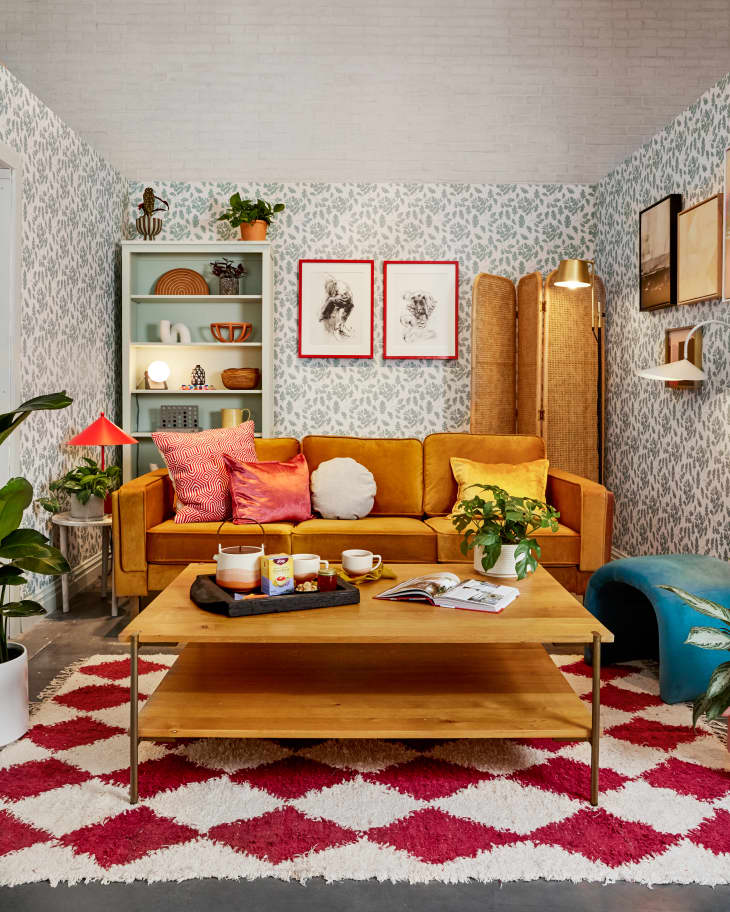 bright happy living space with gold velvet sofa, coffee table with yogi tea setup, patterned wallpaper, red and white rug, shelves with l'objets, plants