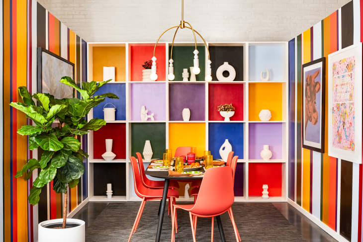 Dining room with multi-colored painted cube shelves, each holding a white object. Bright striped walls, orange dining chairs and colorful tableware