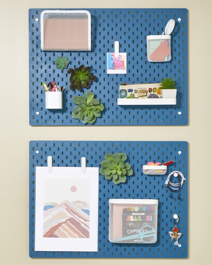 Head on view of two blue pegboards hanging on a beige wall. Each pegboard has an assortment of small plants, pen cups and art.