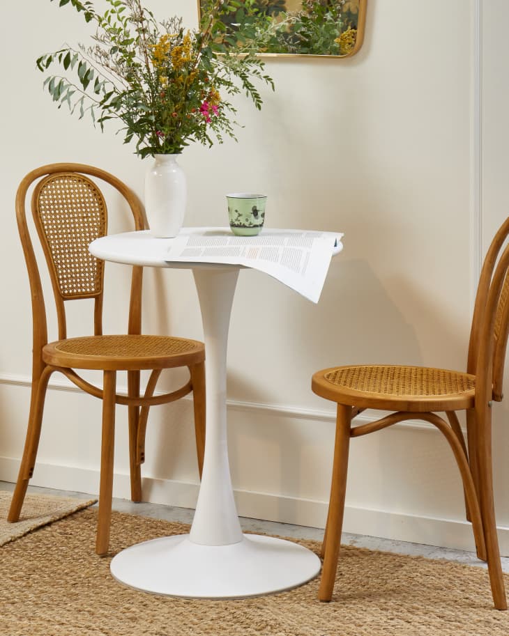 Close up view of two light wood bistro chairs, and a small white tulip table with a white vase full of greenery and flowers.