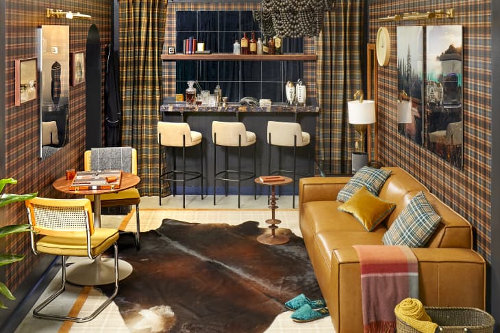 Head on view of a living room with black and yellow plaid wallpaper, a light brown leather couch and a wall of mirrors with a floating bar top and shelves against the wall.