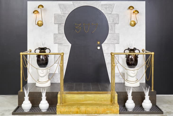 Head on view of a keyhole shaped doorway, with the number 307 written in gold chain on the front.  The door is flanked by 2 bust stools holding black plant urns with white plants in them.