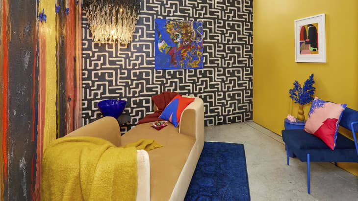 Angled view of a corner of a room with black and white patterned wallpaper on the right wall, and a multicolor painted wall with blue butterflies on the left. In the middle of the room is a neutral colored velvet couch with a yellow throw blanket on it.