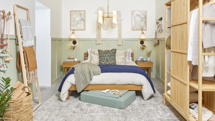 Head on view of a bedroom with white walls, sage wainscoting and a neutral patterned area rug. The bed and bedside tables are a light brown wood and the bedding is beige with a blue throw and a sage green throw and a blue and green decorative throw pillow and theres a green dog bed at the foot of the bed.