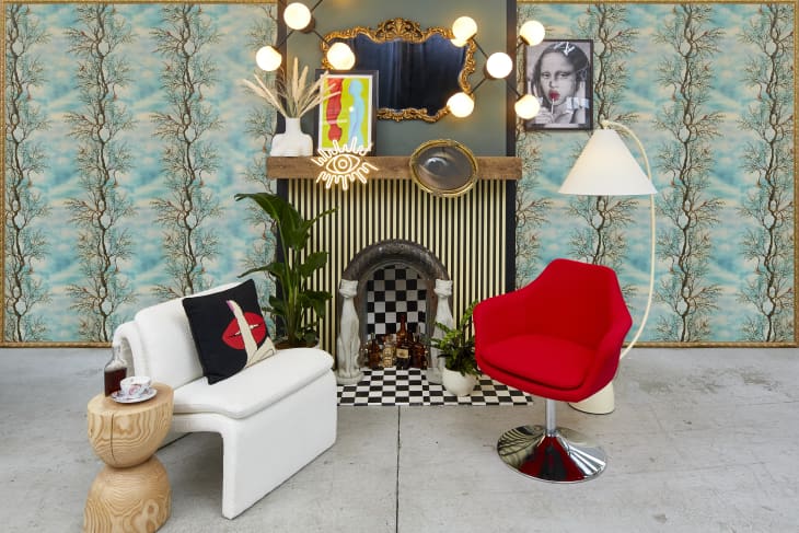 Head on view of a room with a blue and tree printed wallpaper, a striped and checkered fireplace and one white chair and one red swivel chair on either side of the fireplace.