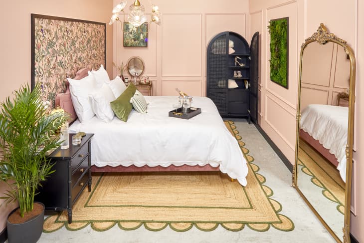 Side view of a pink velvet bed frame with white scalloped duvet. The room has pink walls, a black armoire and side table, and a gold framed standing mirror.